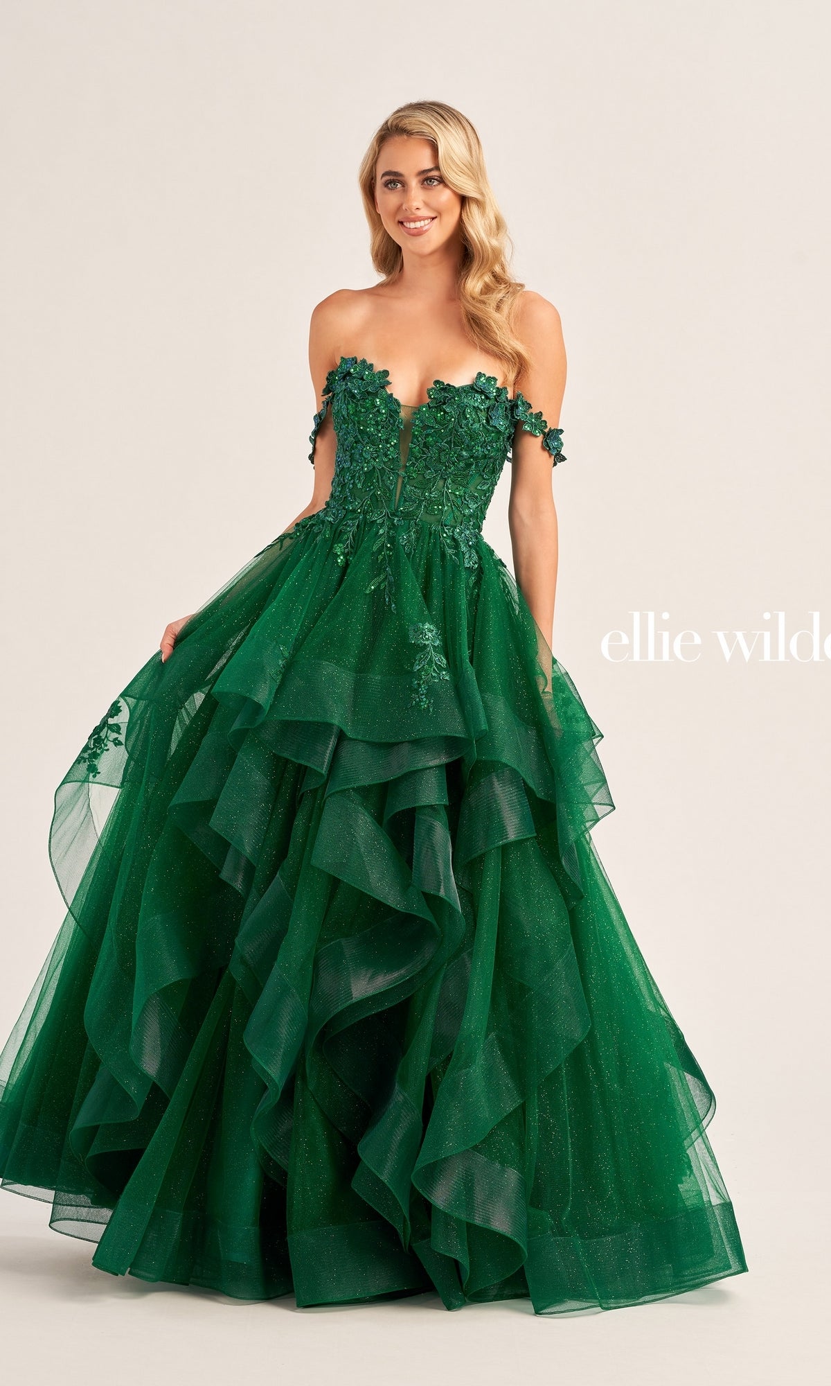 A woman stands against a plain background wearing the Ellie Wilde Long Designer Prom Ball Gown EW35084, a strapless, emerald green glitter tulle dress with tiered layers and floral embellishments.